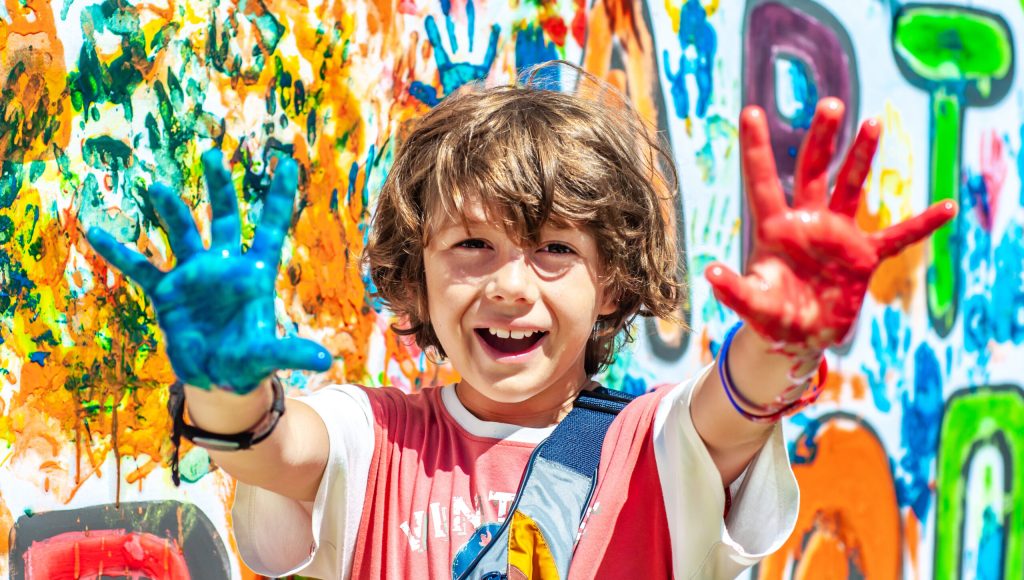 Happy cute boy with painted paint palms outstretched ahead. Happy summer holidays. School education. Children's summer camp outdoors