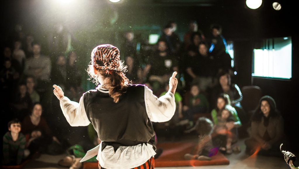 A performing arts entertainer is seen from the back in selective focus, dressed as a pirate on stage during a comedy act with blurry audience at back.