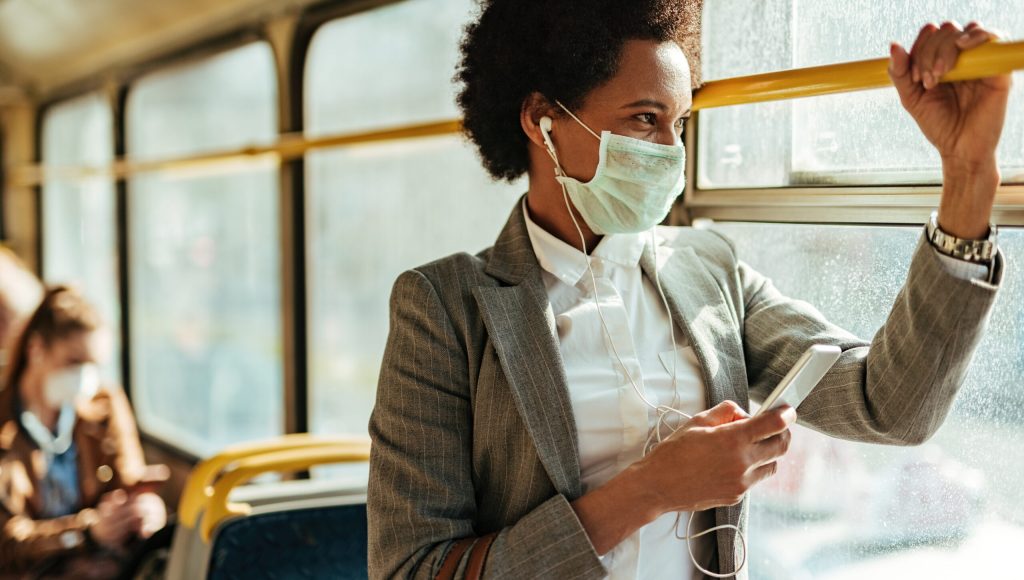 Black businesswoman with protective face mask using smart phone and looking through the window while commuting by bus.
