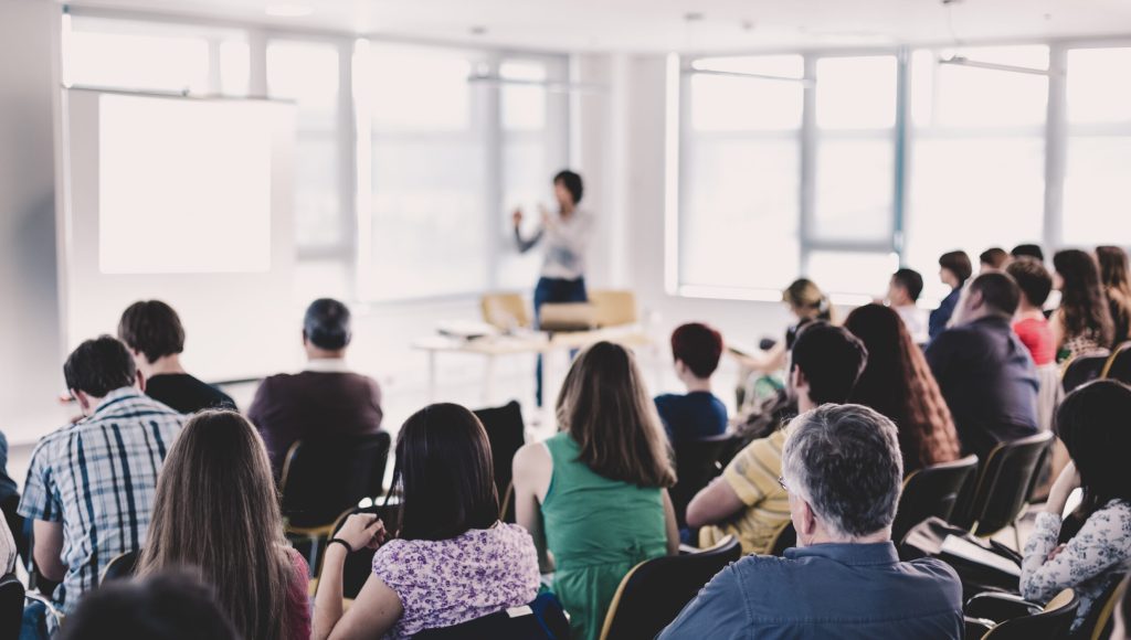 Business and entrepreneurship symposium. Speaker giving a talk at business meeting. Audience in conference hall. Rear view of unrecognized participant in audience.